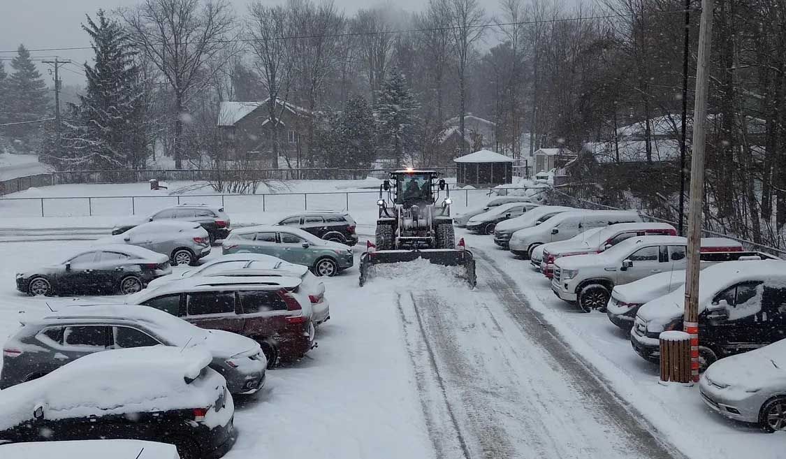 Une déneigeuse dégage un parking enneigé bordé de voitures stationnées.