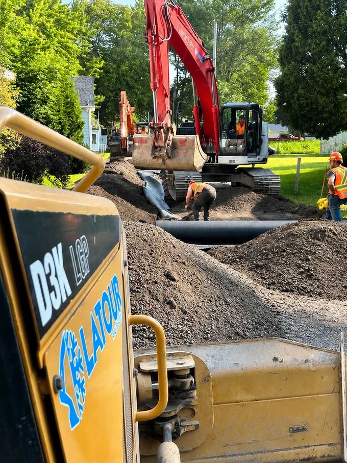 Une équipe de construction creuse une tranchée avec une excavatrice rouge et de la terre boueuse sur un chantier en bord de route