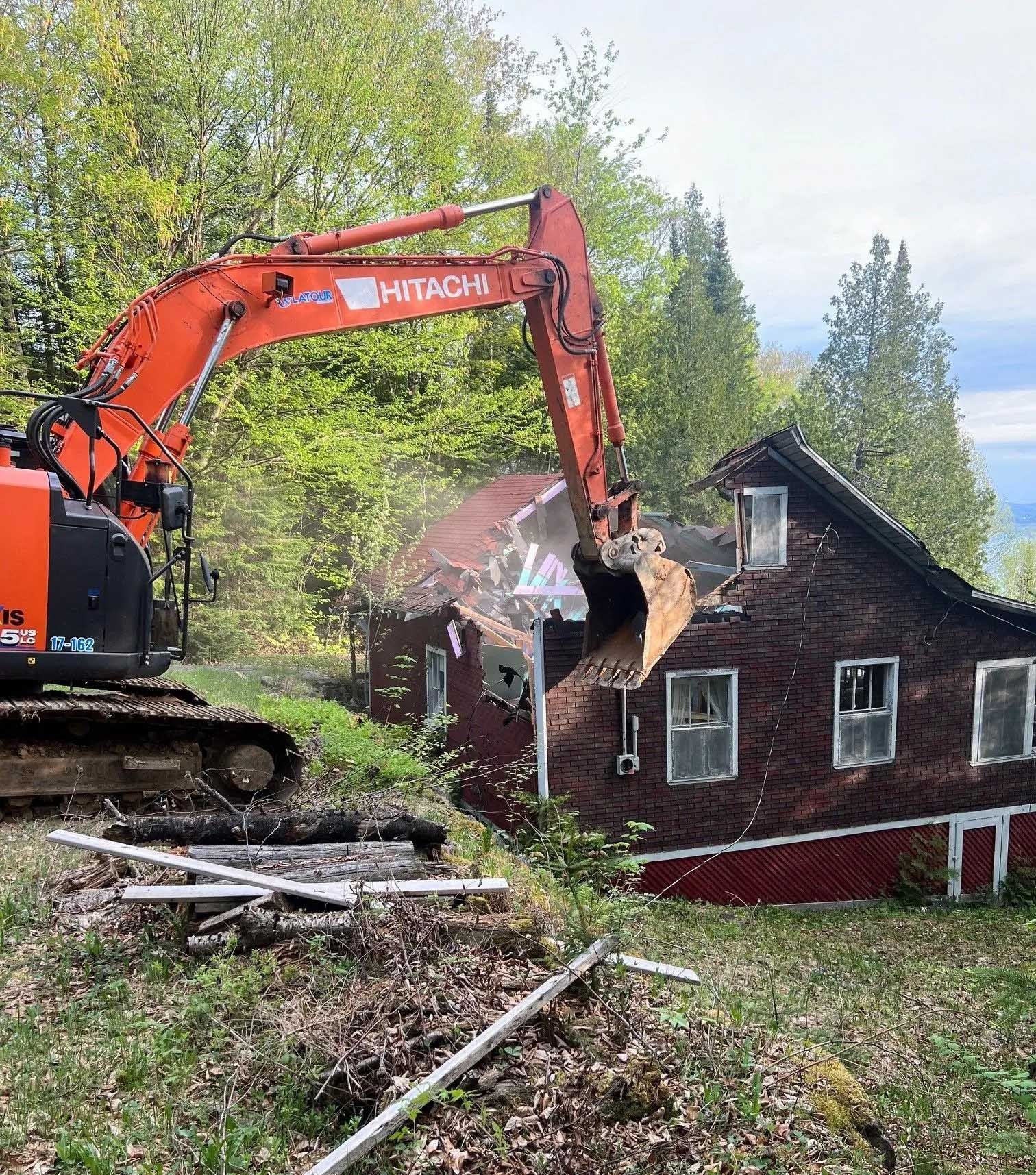 Une pelleteuse orange démolit une maison en bois sombre sur une colline boisée.