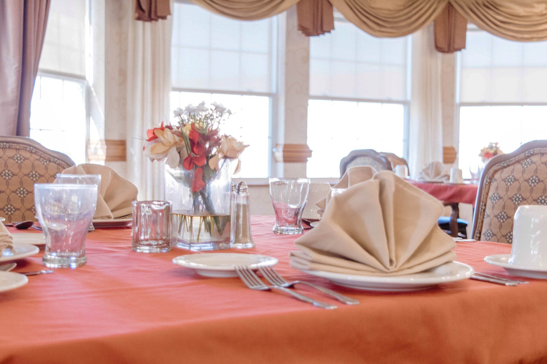 A dining table set for a meal, with a centerpiece of flowers, and large windows.