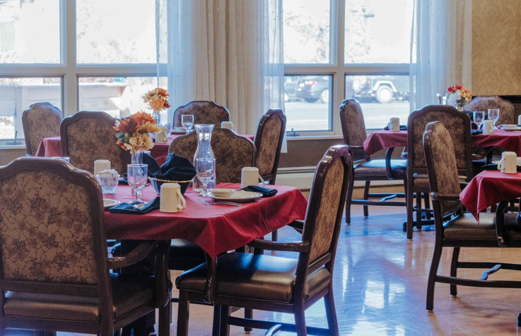 Dining room with tables set for a meal. Red tablecloths, ornate chairs, and a large window in the background.