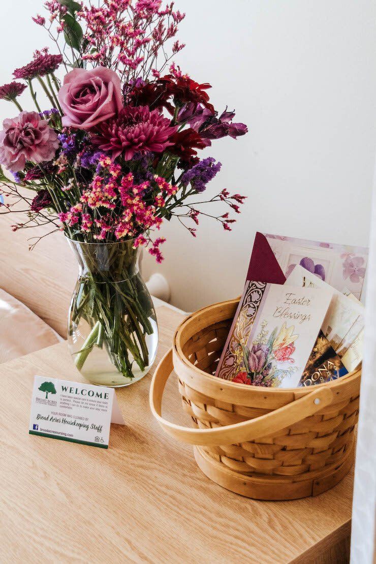 A bouquet of purple flowers in a vase and a basket of cards on a light-colored wood nightstand.