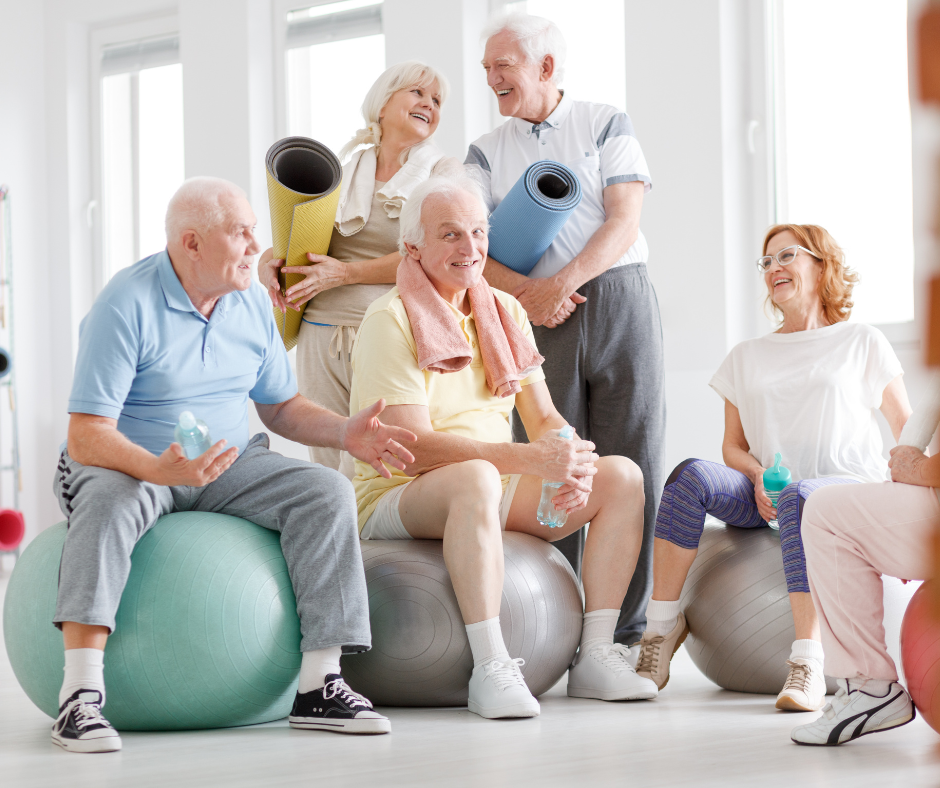 Group of seniors in a gym, sitting on exercise balls, smiling and talking; holding water bottles and yoga mats.