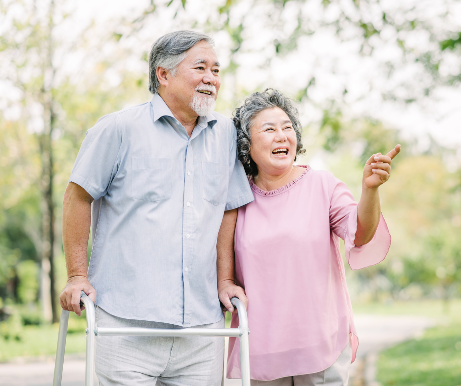 Elderly couple smiles in a park. Man uses a walker, woman points. Outdoors, daytime.