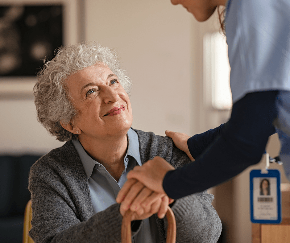 Older woman smiles, looking up at a caregiver. Caregiver’s hand on her shoulder. Indoor setting.