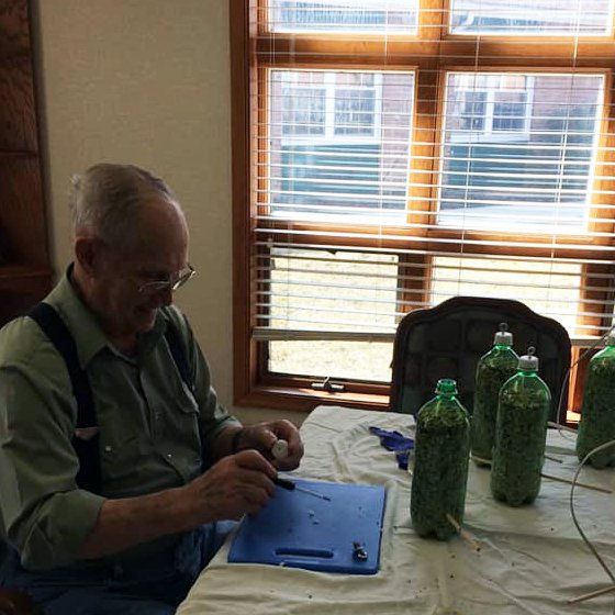 An elderly man works on craft project at a table near a window. He is using tools and surrounded by bottles.