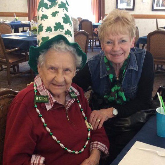 Two women smiling, one wearing a St. Patrick's Day hat and necklace. Indoors, tables with blue tablecloths.