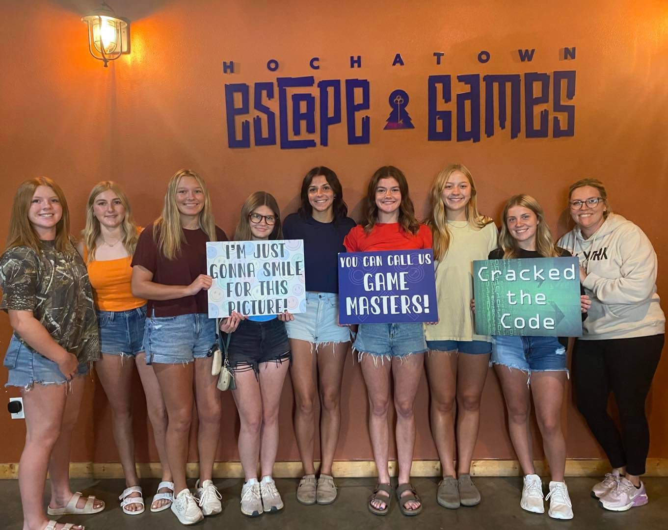 Group holding coding-themed signs in front of a Hochatown Escape & Games wall mural.
