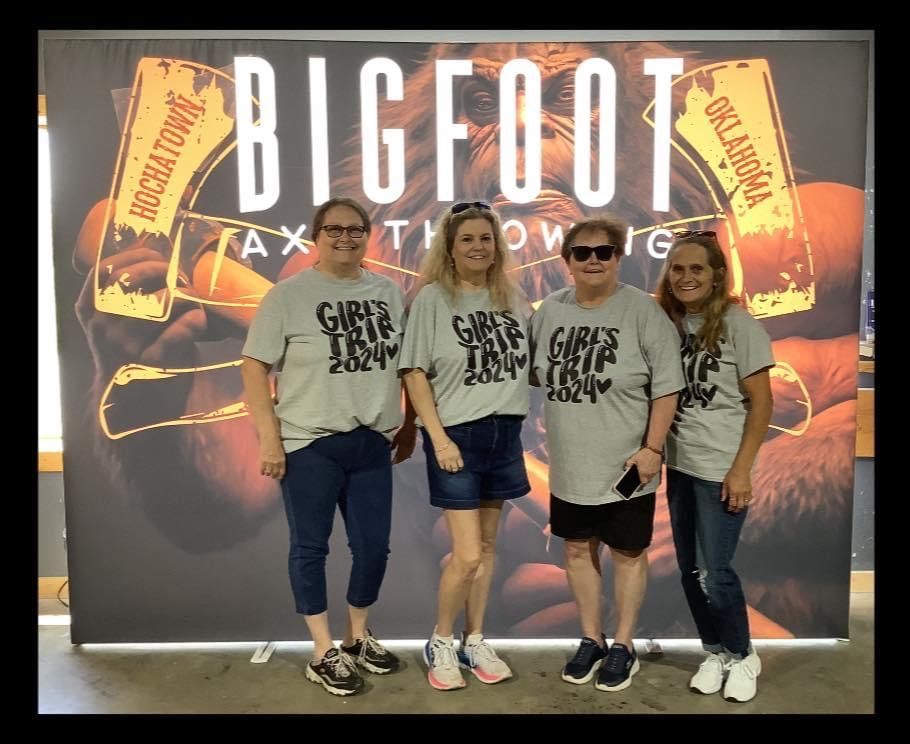 Four people in matching gray shirts pose in front of a Bigfoot-themed backdrop.