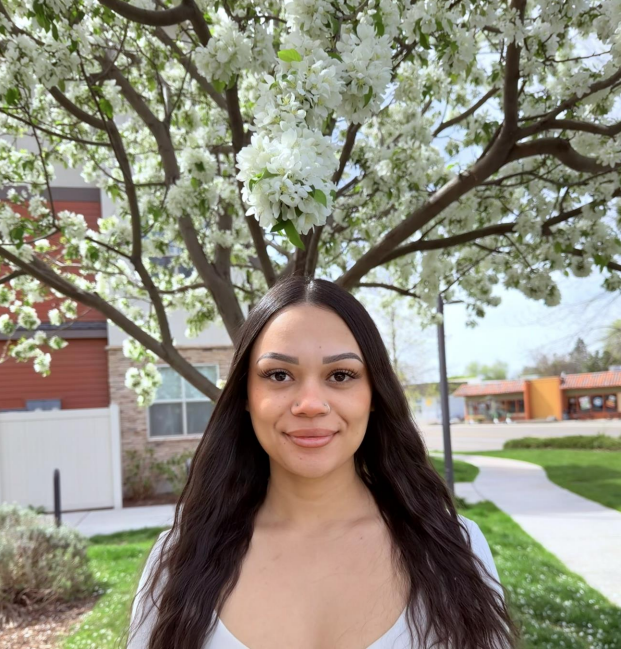 Smiling woman in a brown polka-dot blouse standing outdoors among blooming white flowers.