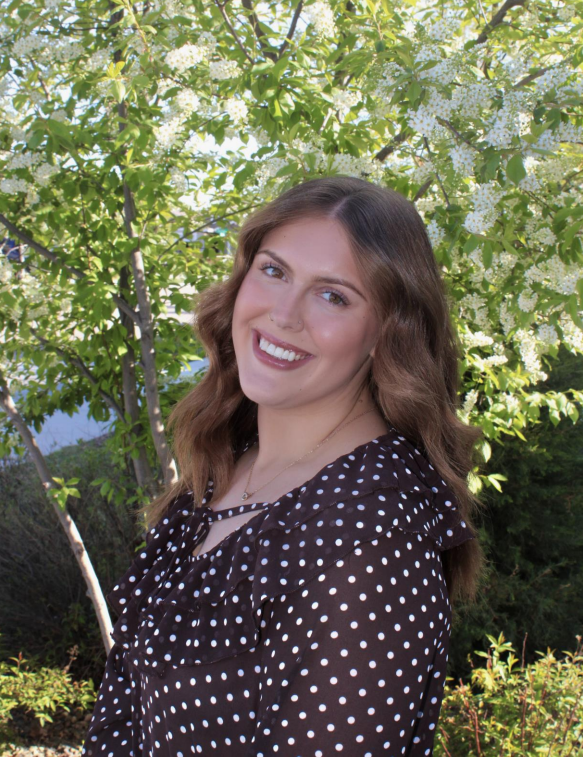 Smiling woman in a black polka dot dress standing outdoors among green trees and sunlight