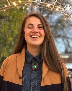 A woman wearing a denim shirt and a brown jacket is smiling for the camera.