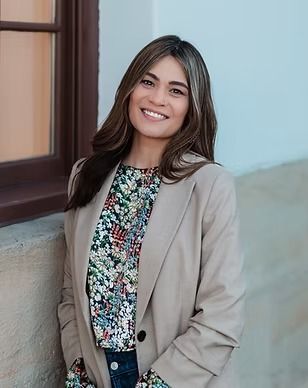 A woman is standing in front of a window wearing a blazer and a floral shirt.