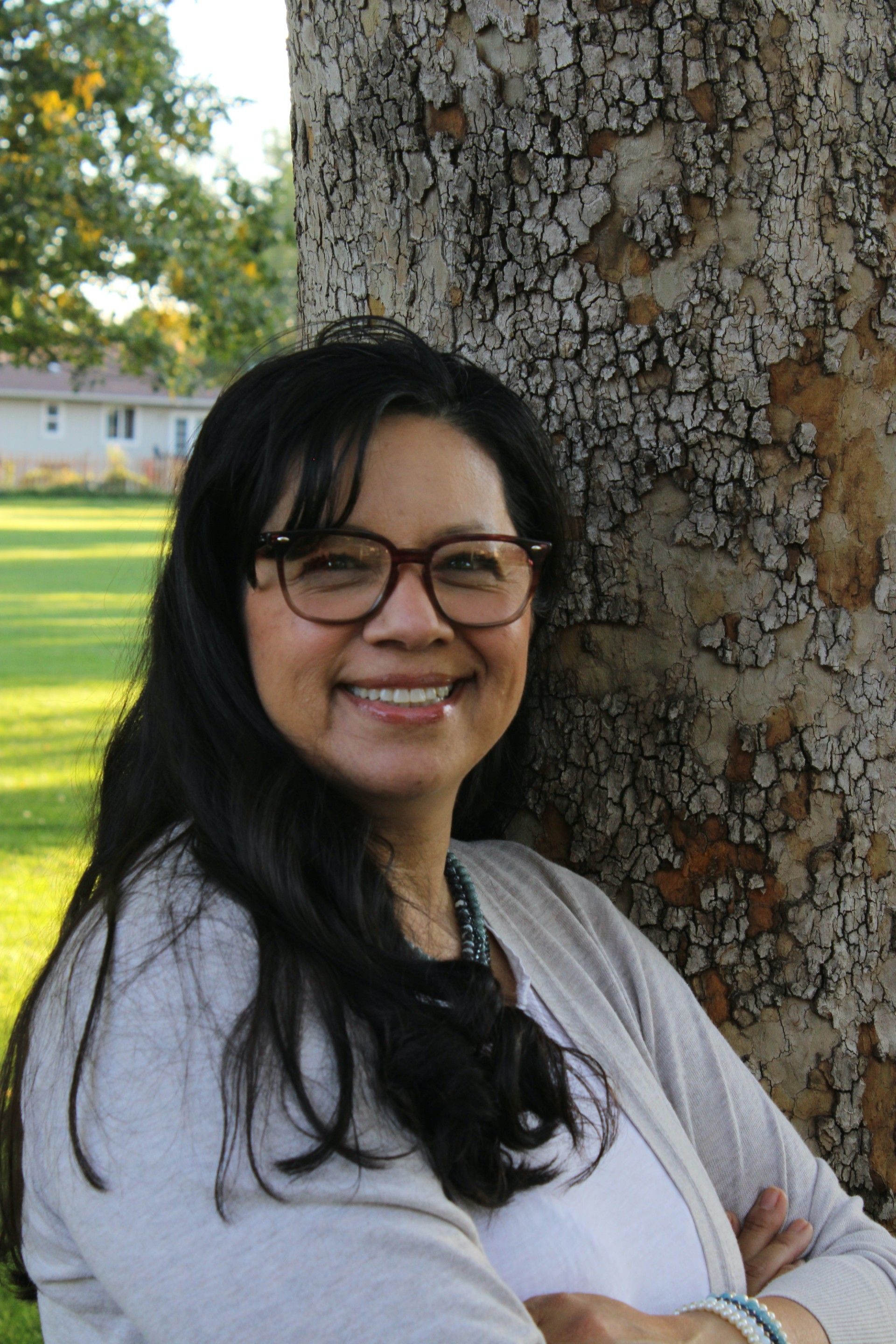 Woman with glasses smiles, leaning against a tree, arms crossed, wearing a light sweater.