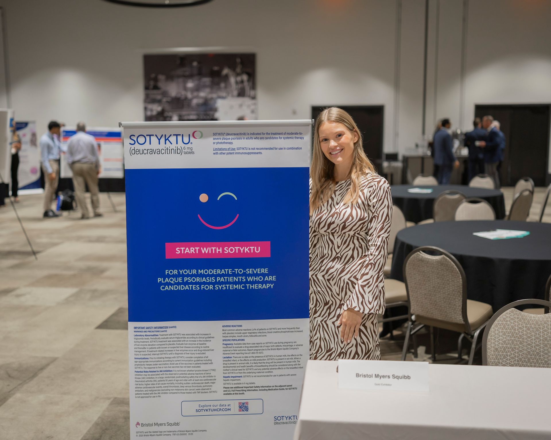 Woman standing beside a blue informational poster at a conference booth in a meeting room.