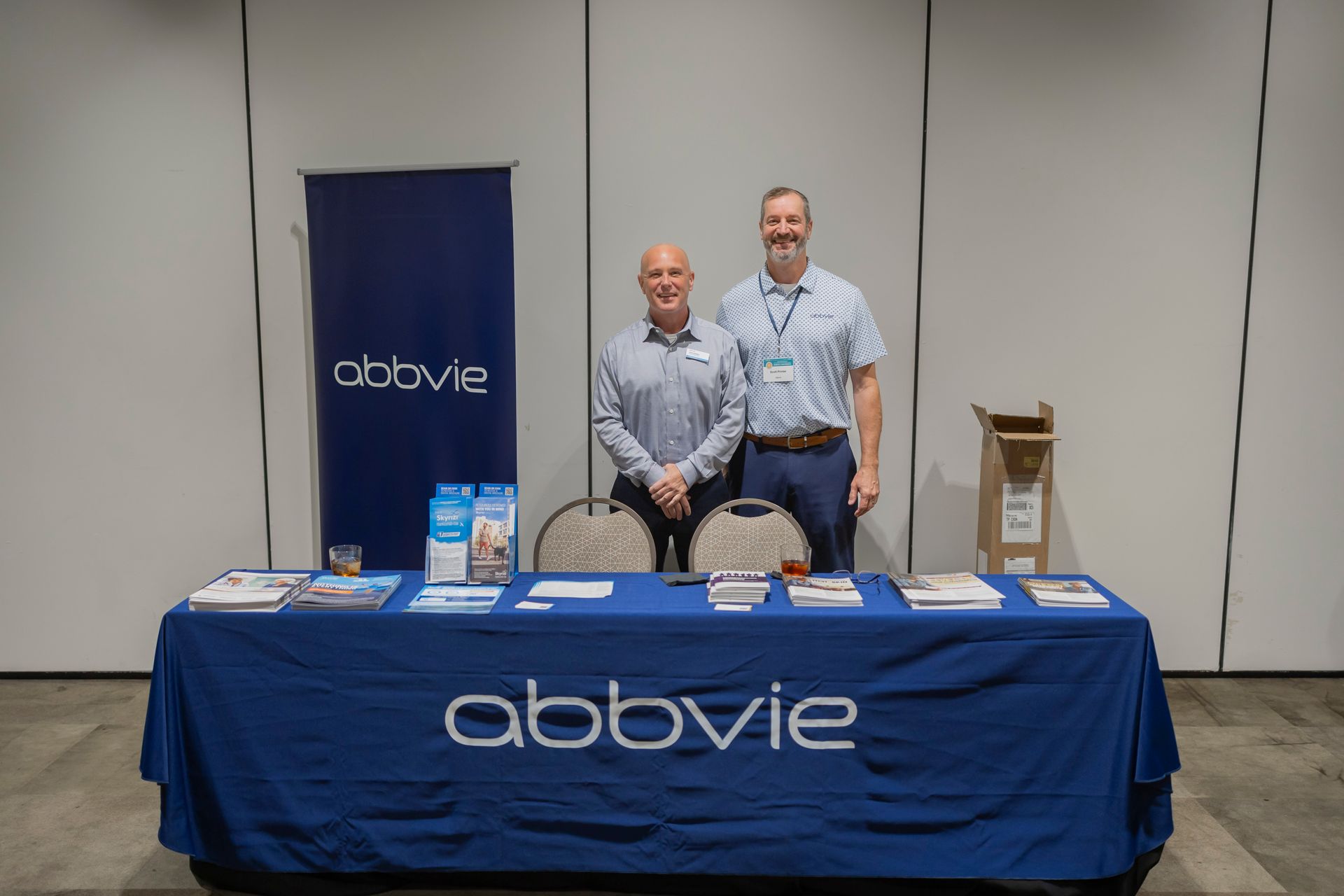 Two people at an AbbVie booth with a blue tablecloth and display items at a conference.