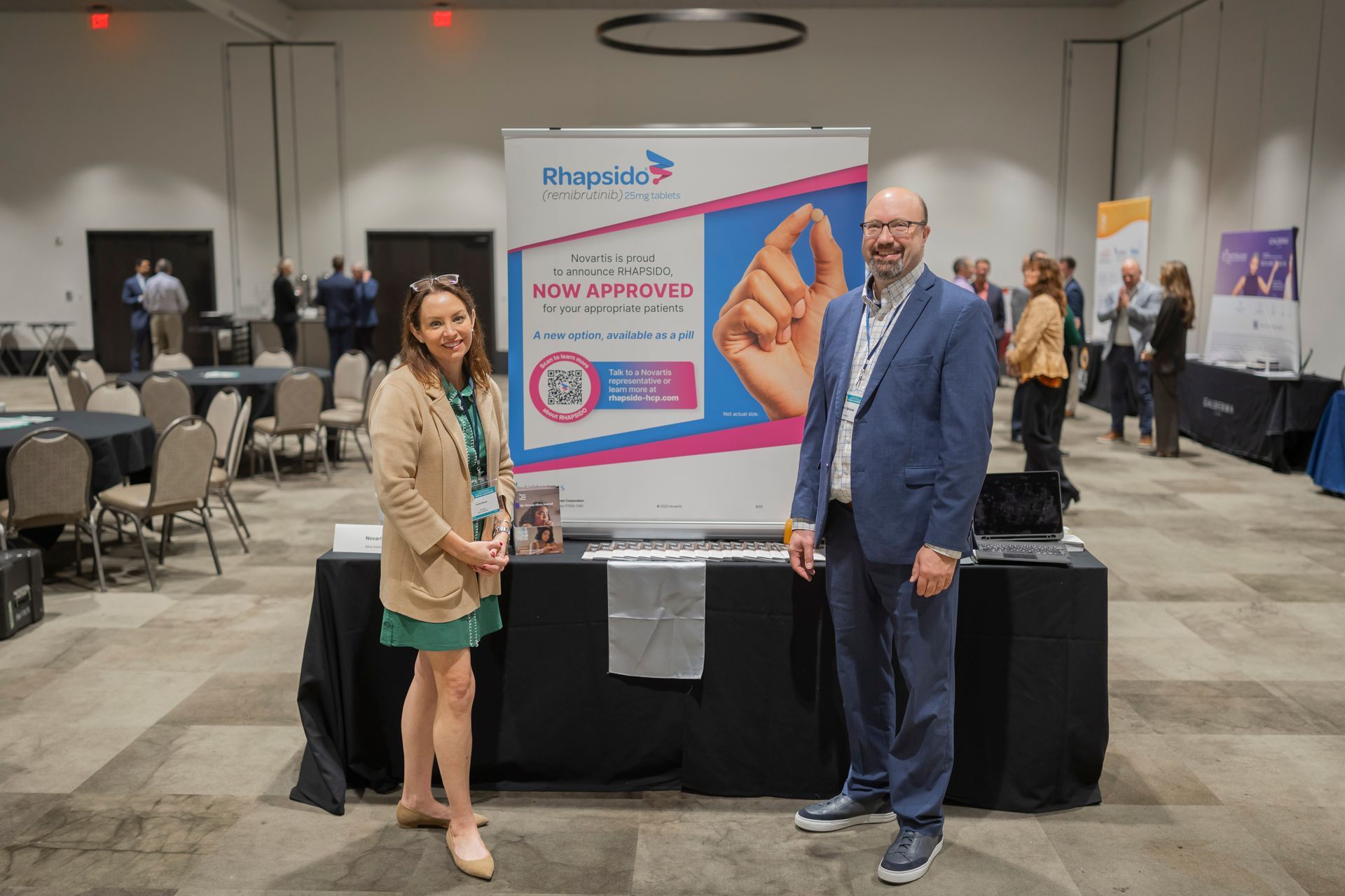Two people posing beside a large poster at a conference booth in a convention hall