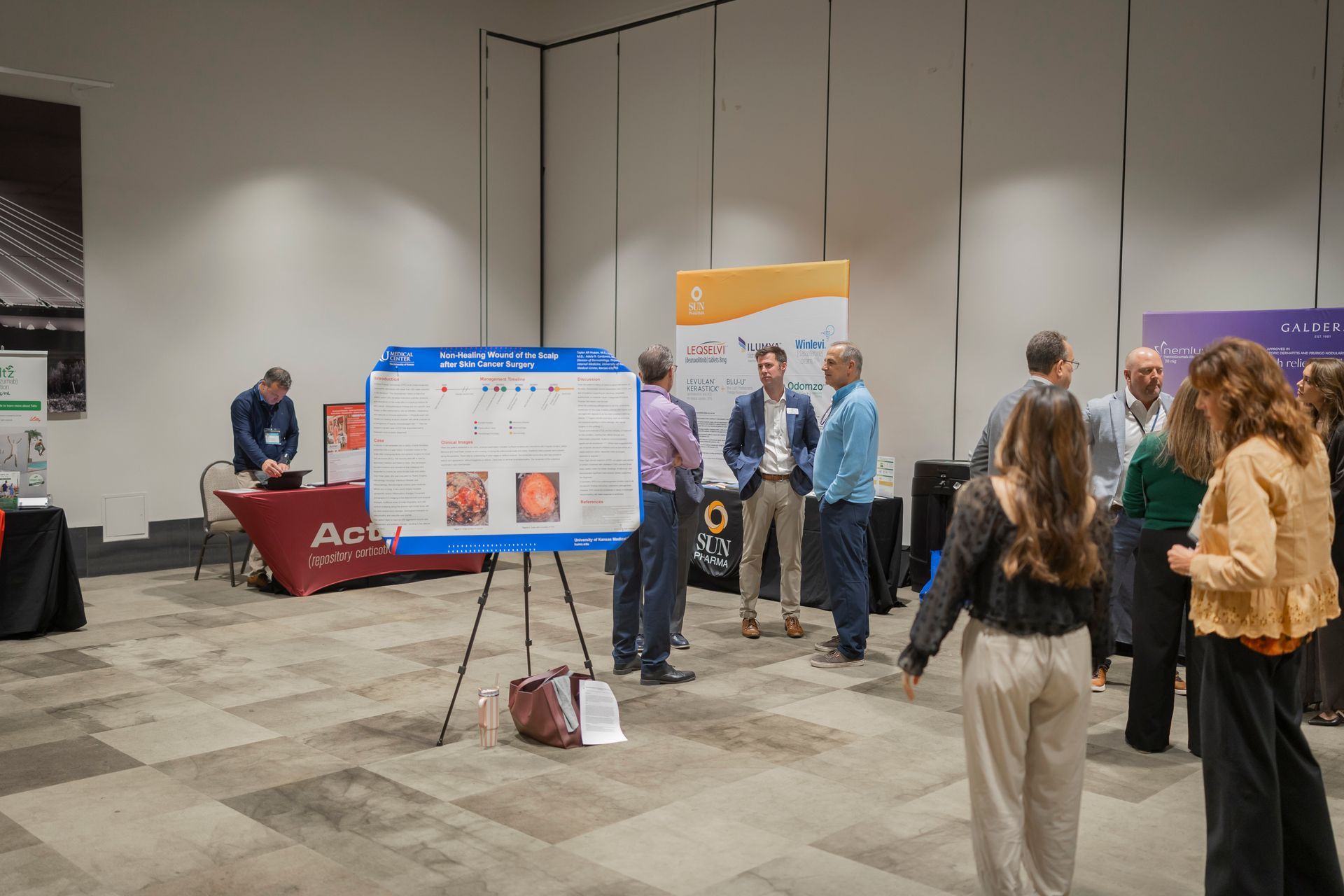 Conference expo hall with booths, a poster display, and small groups of people talking and walking