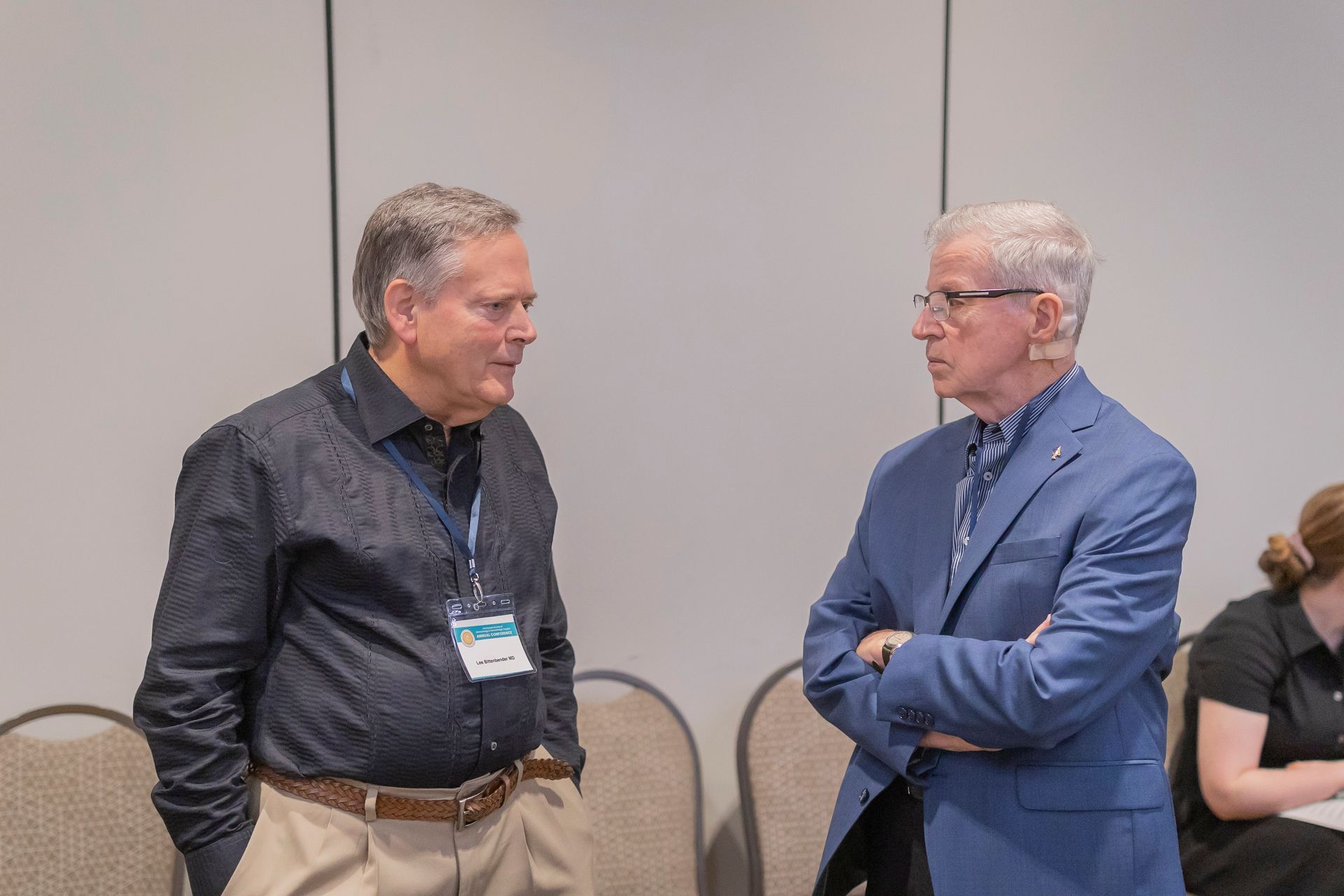 Two men in a conference room talking, one in a dark shirt and one in a blue blazer.