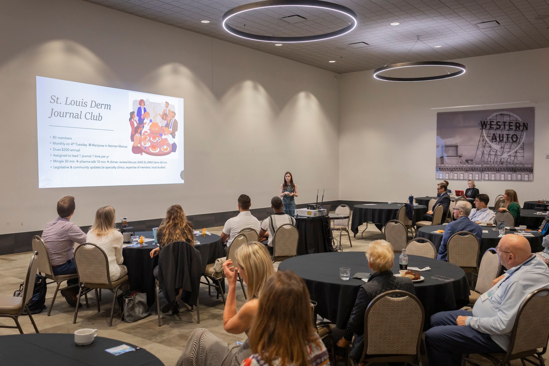Conference room presentation with attendees seated at round tables watching a slide on a projector screen