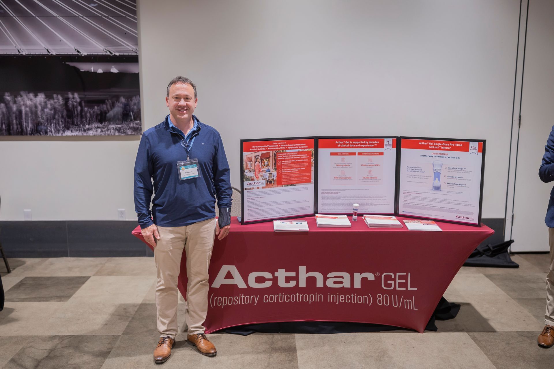 Man standing beside an AcharGBR display table with posters in a conference hall