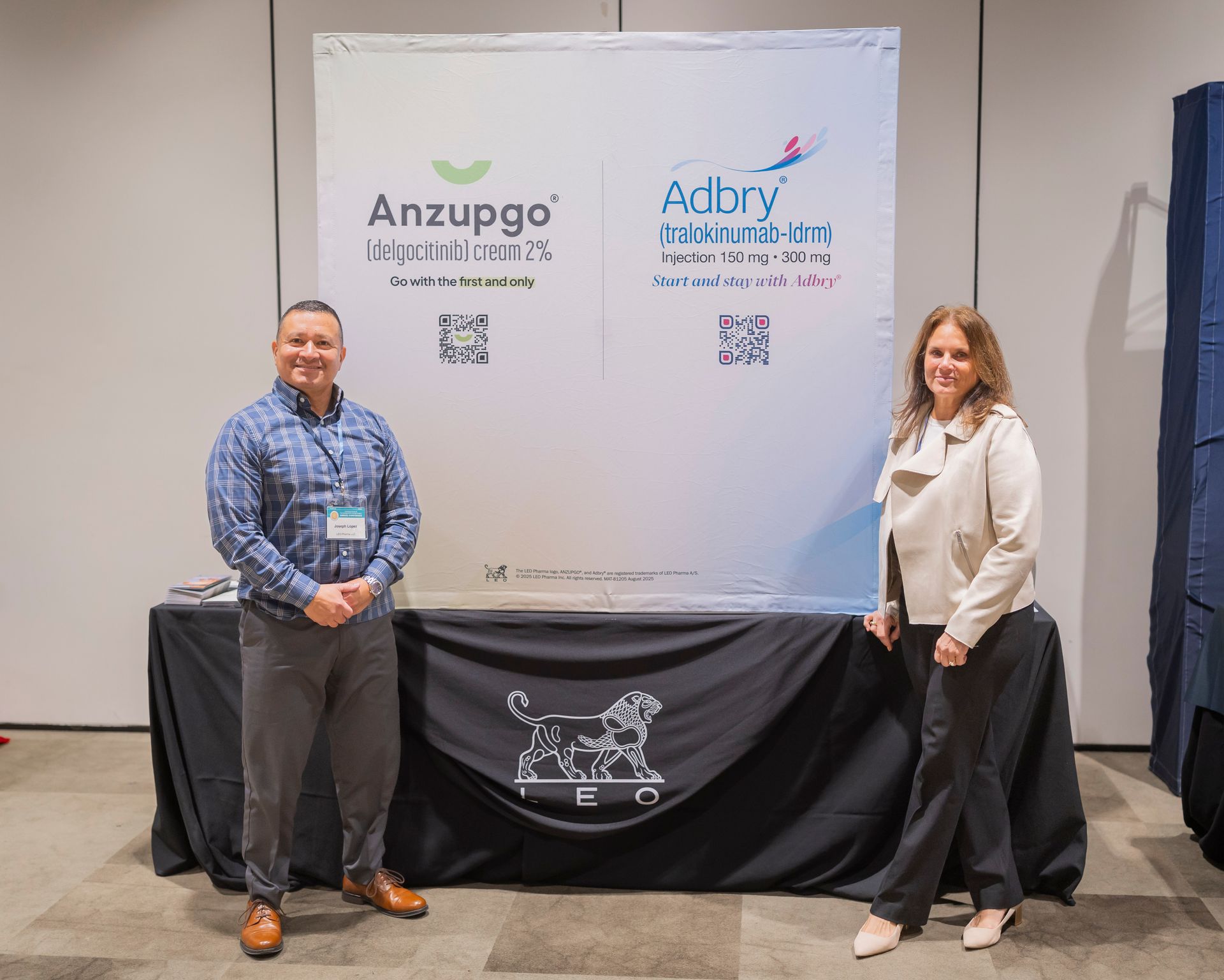 Two people stand beside a branded banner at an event booth, with black tablecloth and white backdrop.