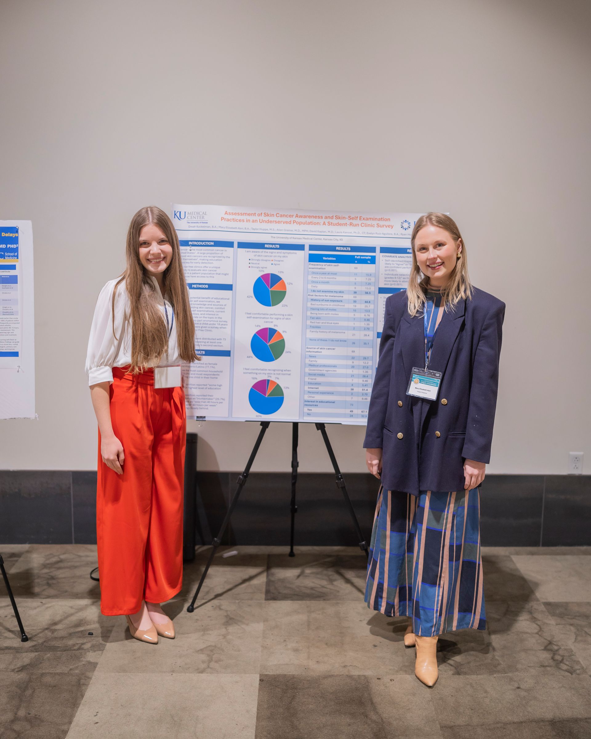 Two women standing beside a poster board with pie charts at a conference display.