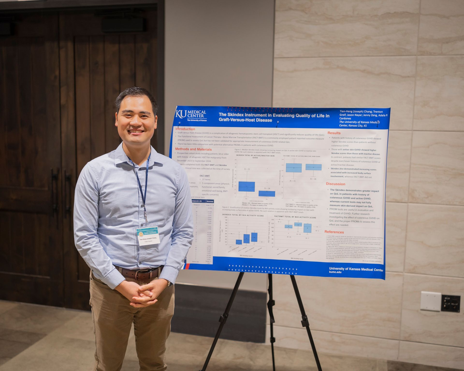 Man standing beside a large blue presentation poster on an easel in a conference hallway.