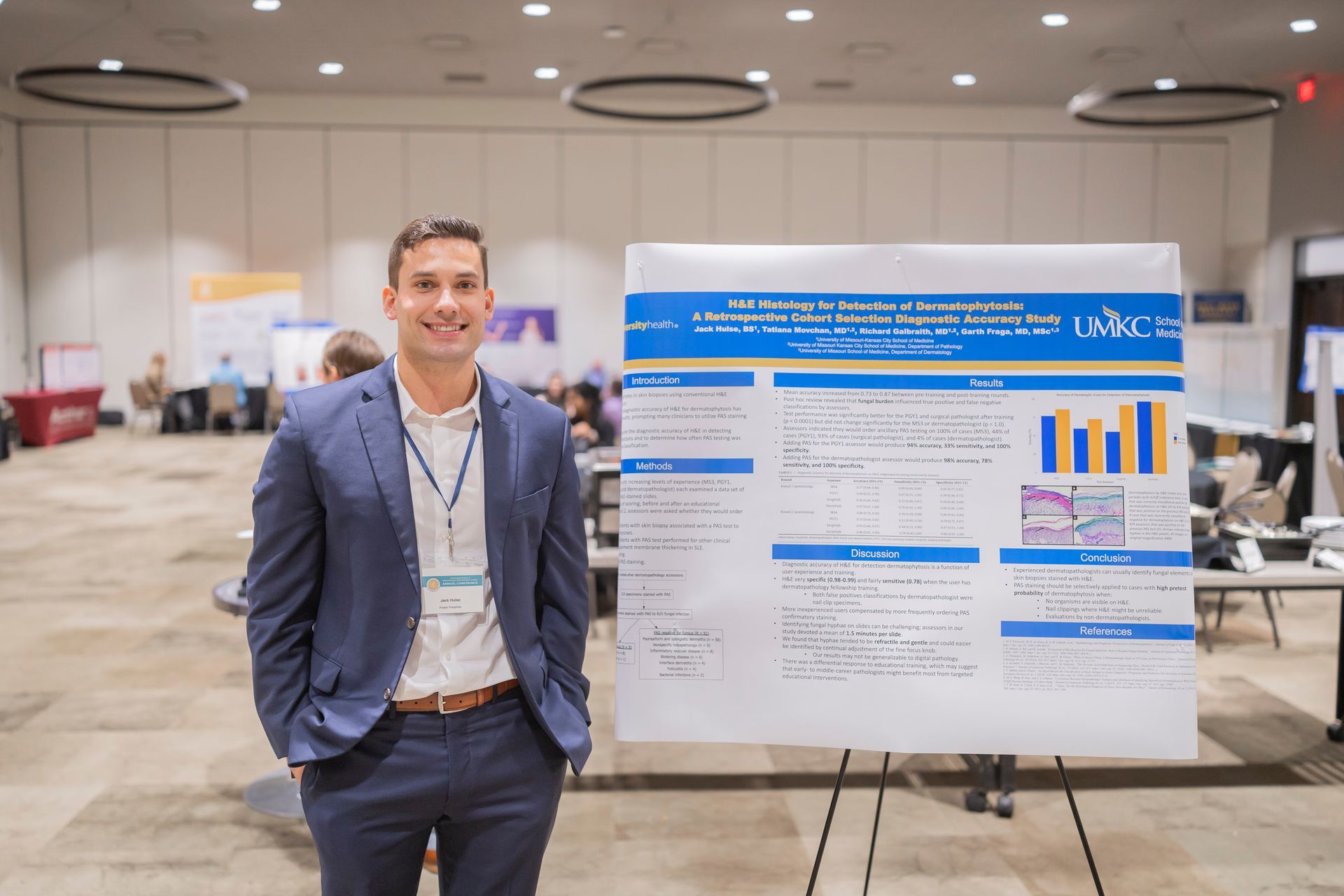 Man in a suit stands beside a conference poster board in a bright convention hall.