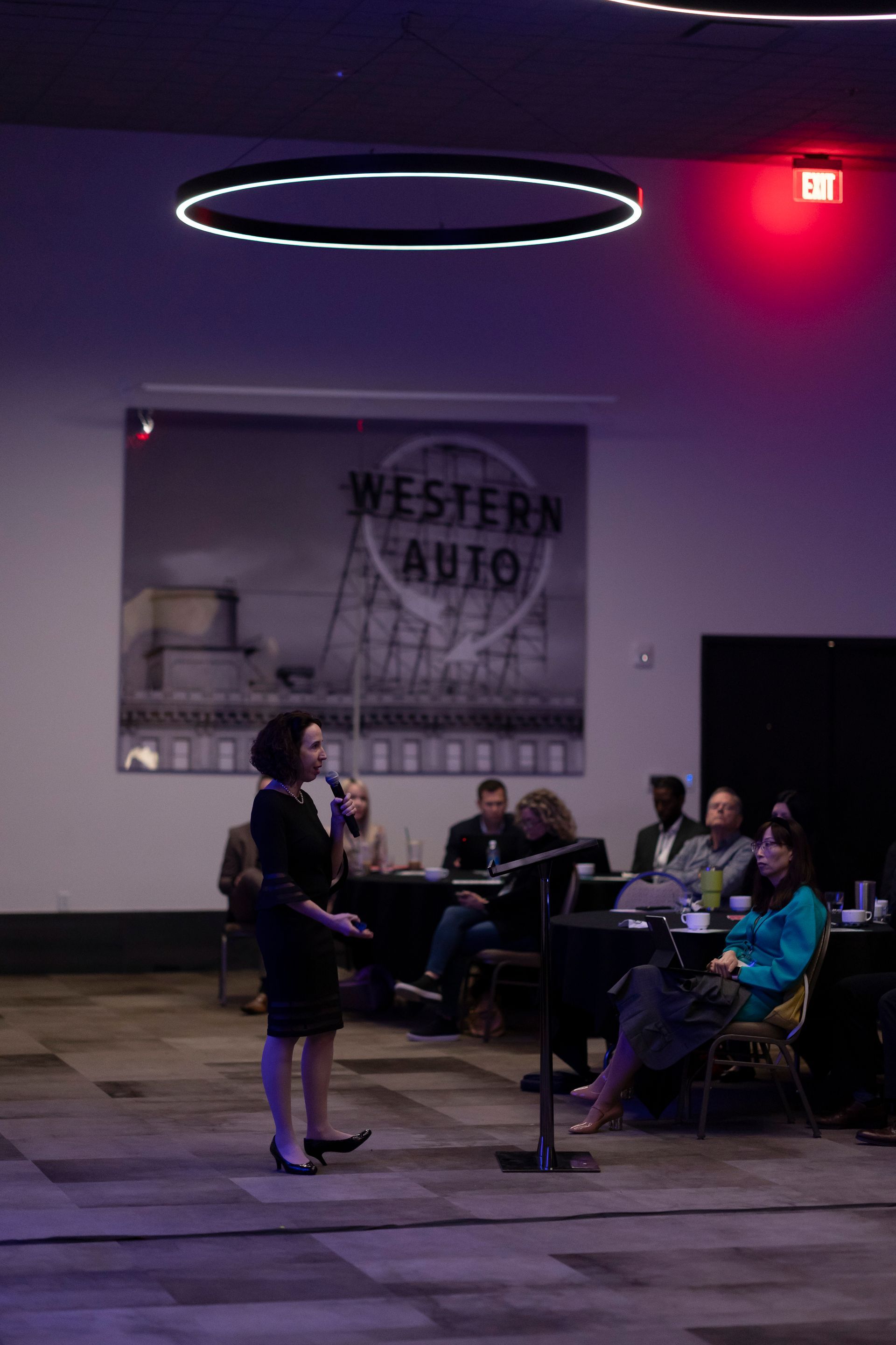A woman is standing in front of a microphone in a room with people sitting at tables.