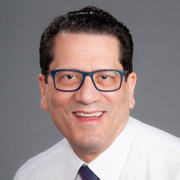 A man wearing glasses and a white shirt and tie is smiling.