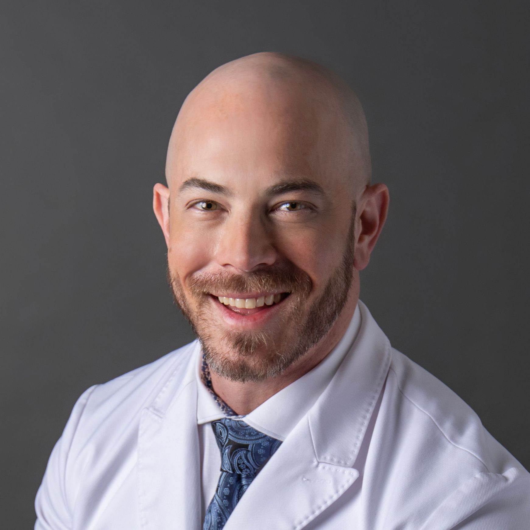 A bald man with a beard wearing a white coat and tie is smiling for the camera.