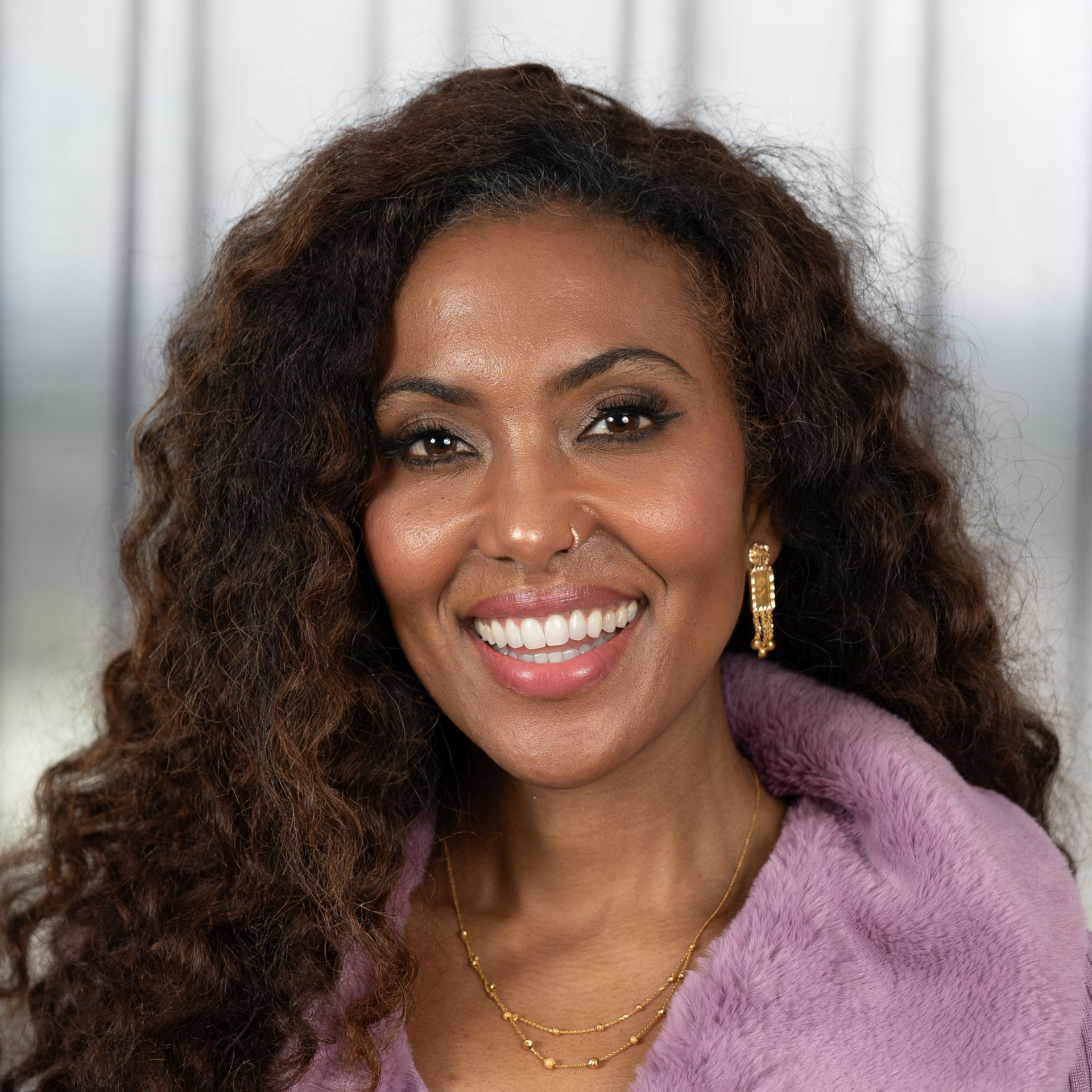 Smiling woman with curly hair wearing a lavender coat and gold jewelry against a soft gray background