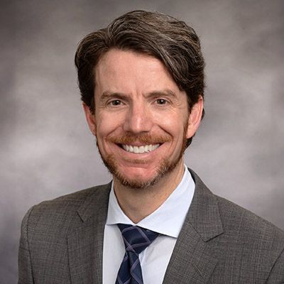 Headshot of a smiling man in a gray suit and blue tie against a neutral studio background