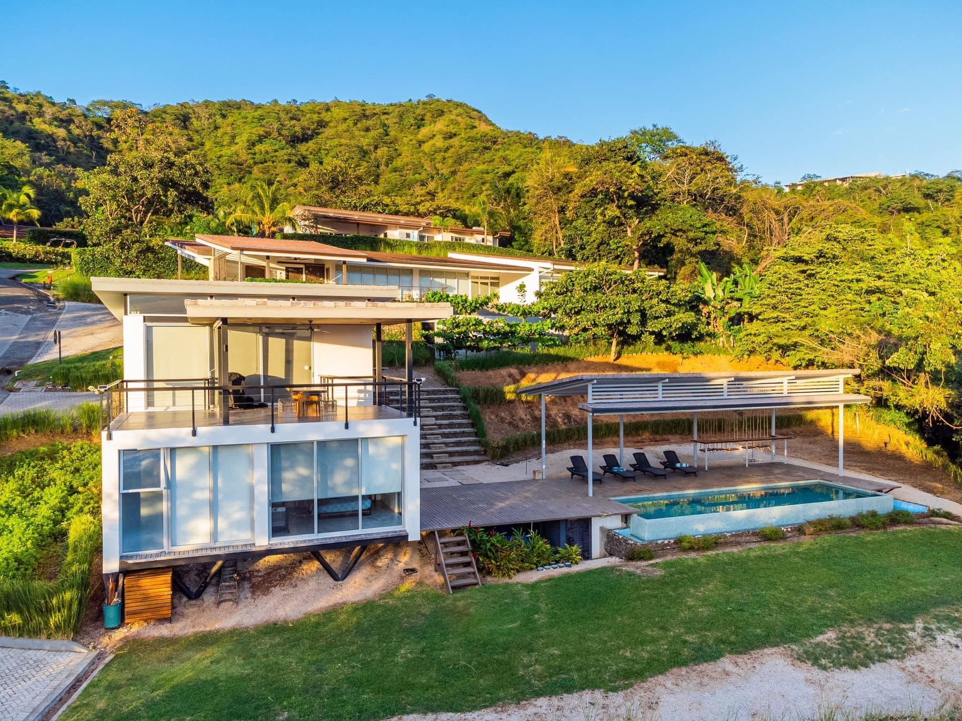Modern white homes with a pool on a grassy hill overlooking a lush green forest, under a blue sky.