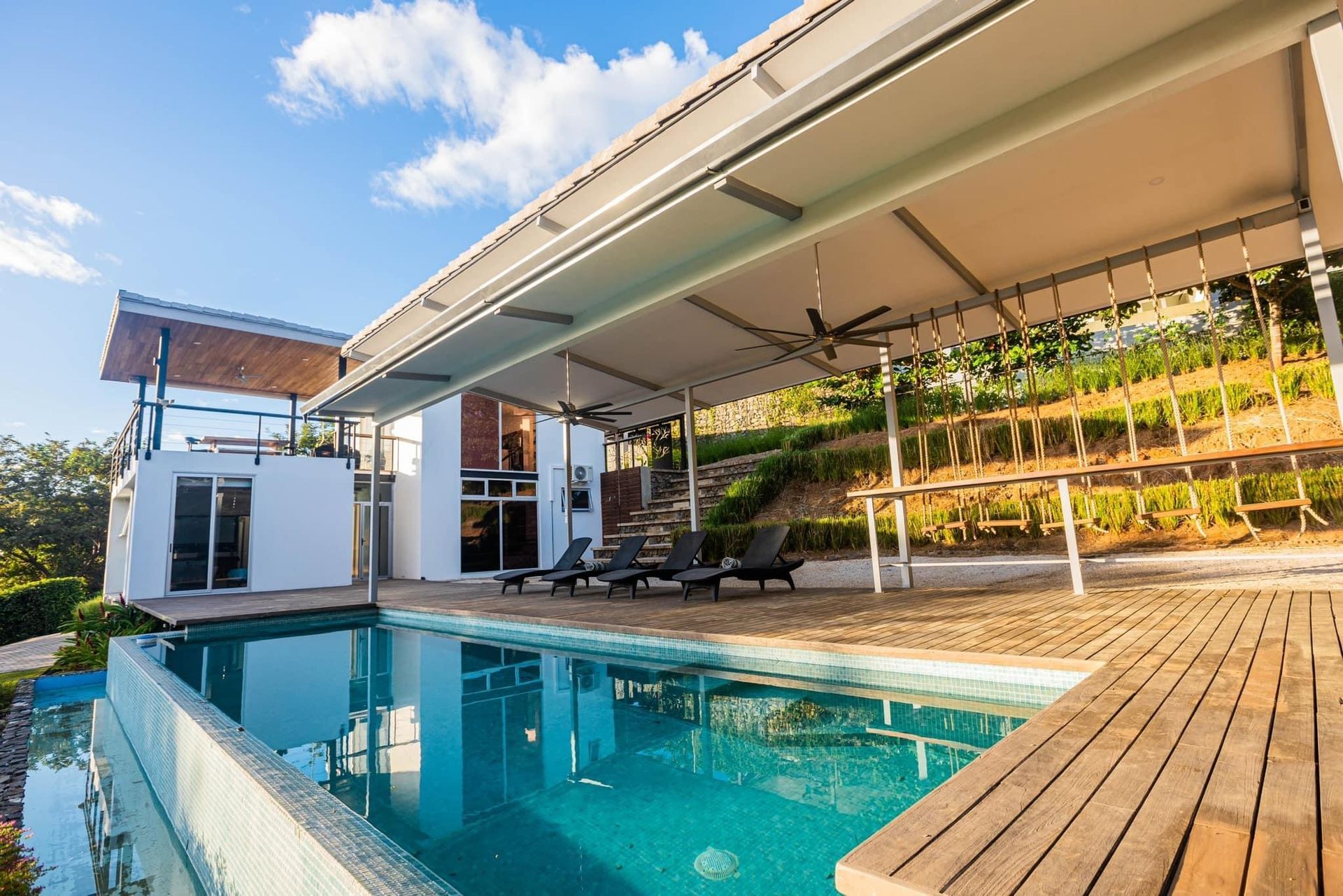 Luxury home with infinity pool, shaded patio, and hillside landscaping. Sunny day.
