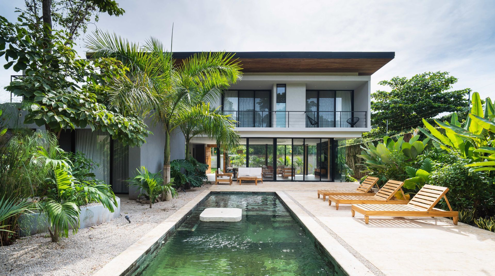 Modern house with a pool and lounge chairs surrounded by lush greenery under a cloudy sky.