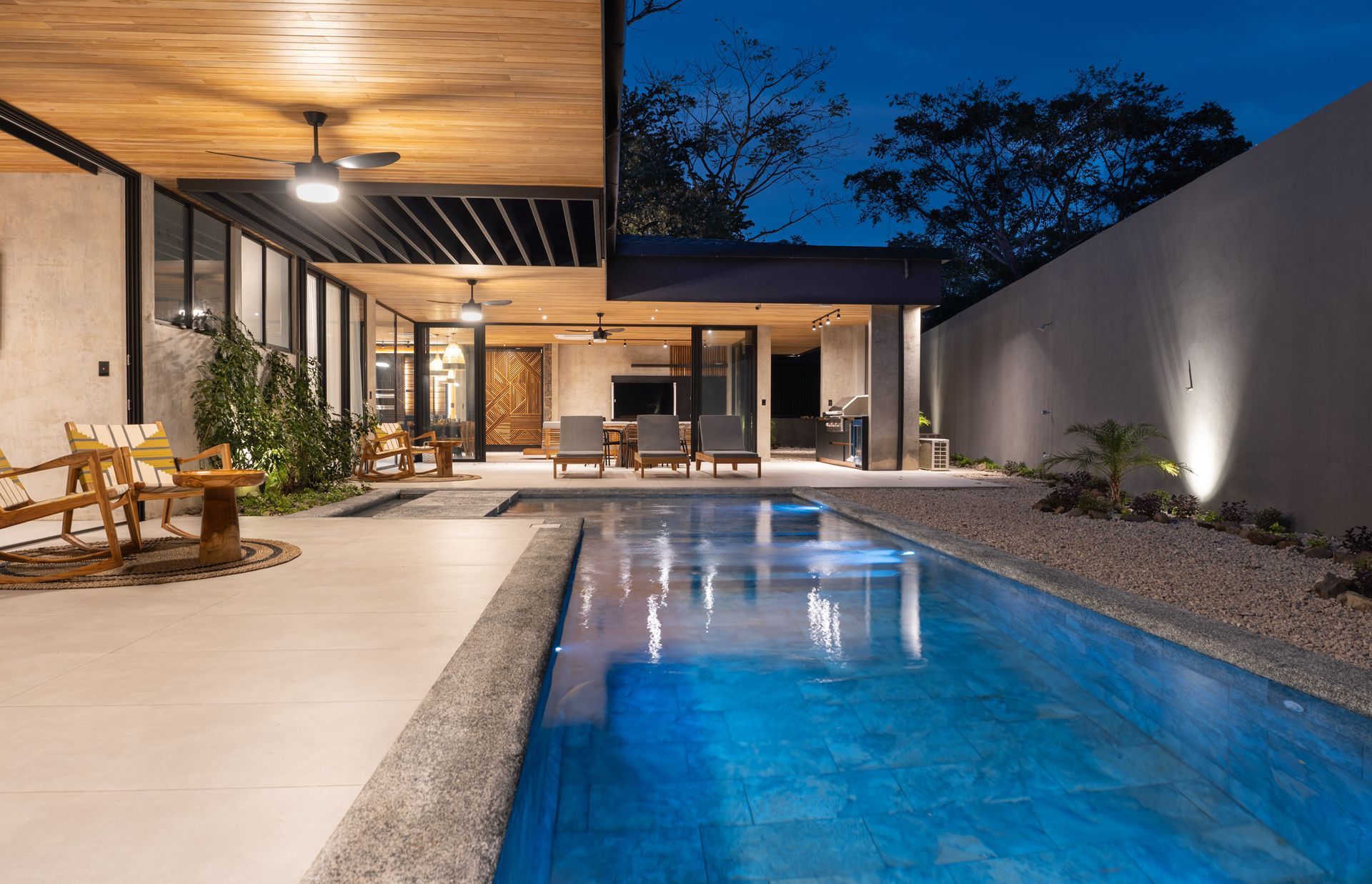 Pool at dusk, modern home. Wooden ceiling, lounge chairs, and tall wall.