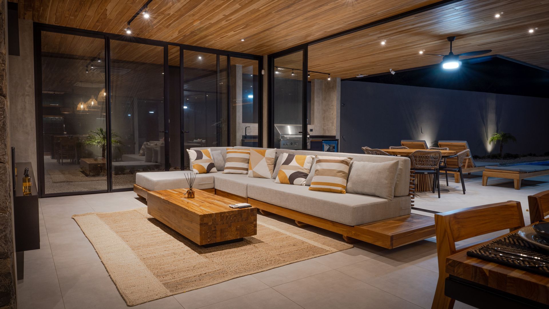 Lounge area with a large gray sectional sofa, wooden coffee table, and woven rug. Large glass windows and a dark setting.