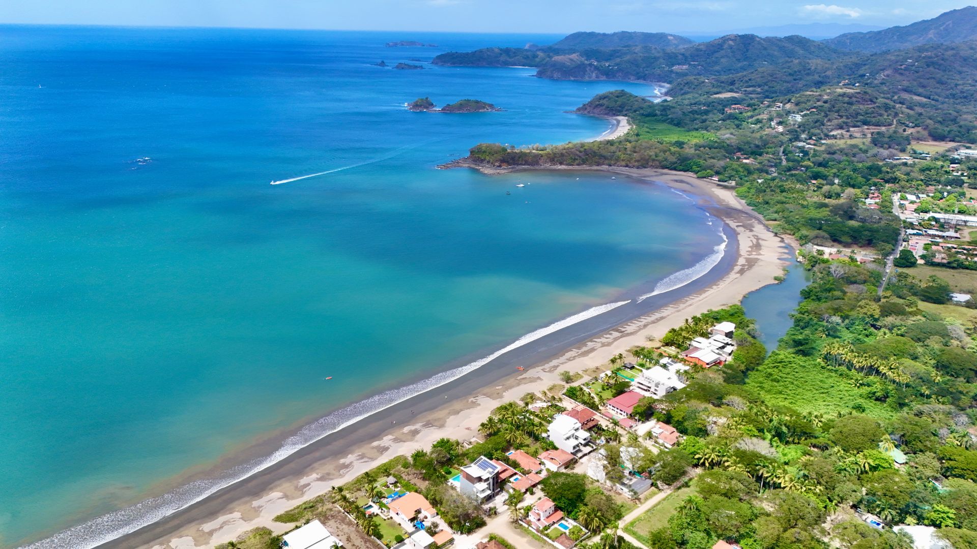 Coastal aerial view: blue ocean, sandy beach, green trees, white buildings along the shore.