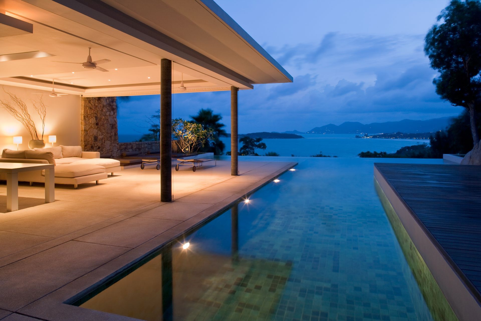 Luxury villa with an infinity pool overlooking the ocean at dusk. Lit interior with open living space.