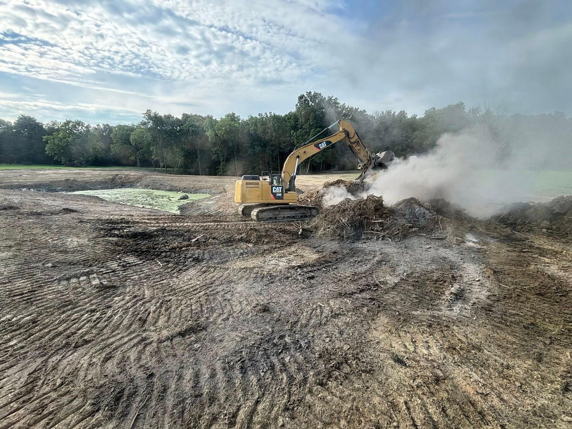 Yellow excavator working on a dirt field, smoke rising. Cloudy sky, trees in background.