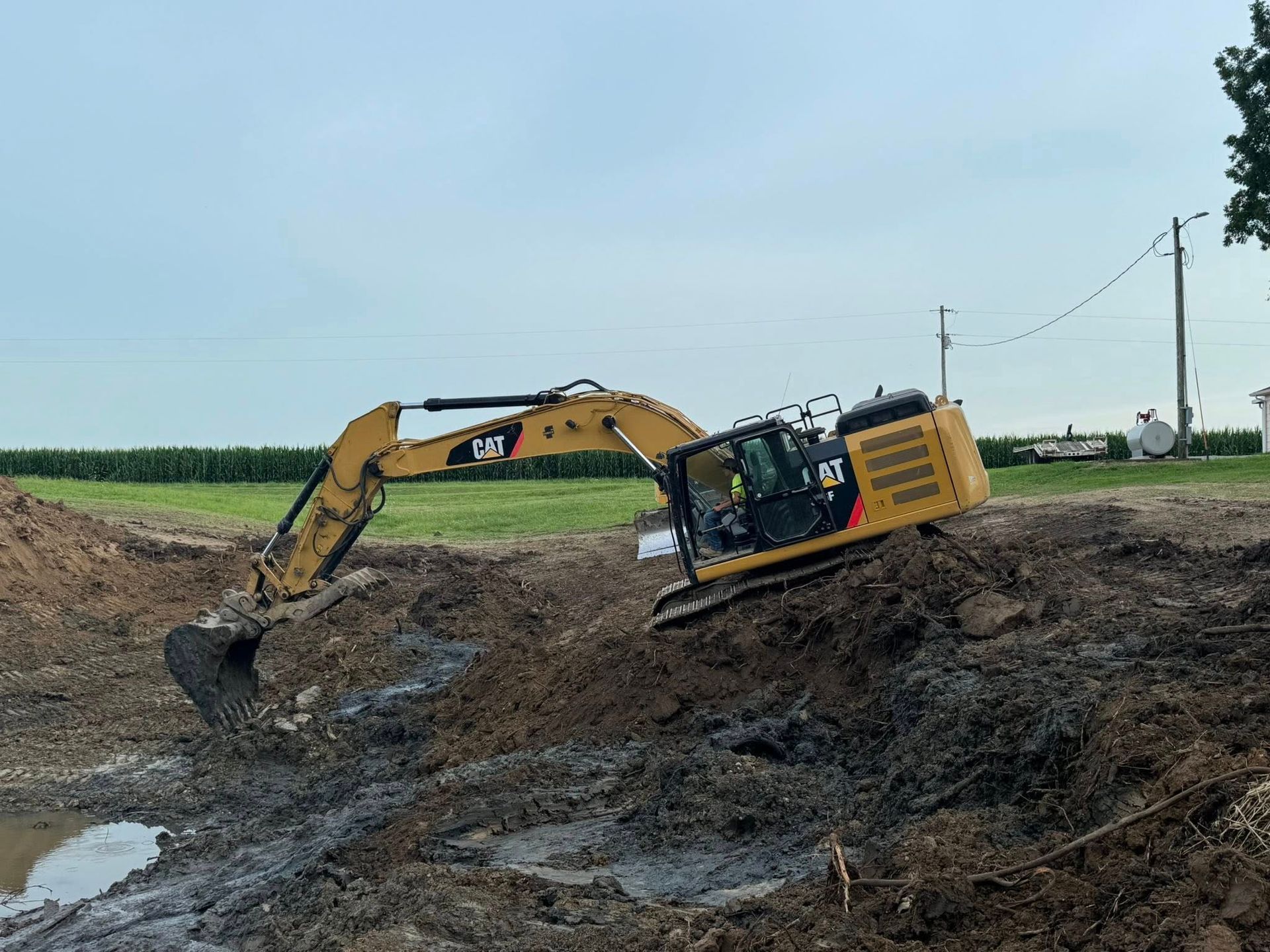 Yellow excavator digging in muddy earth near a field and utility poles.