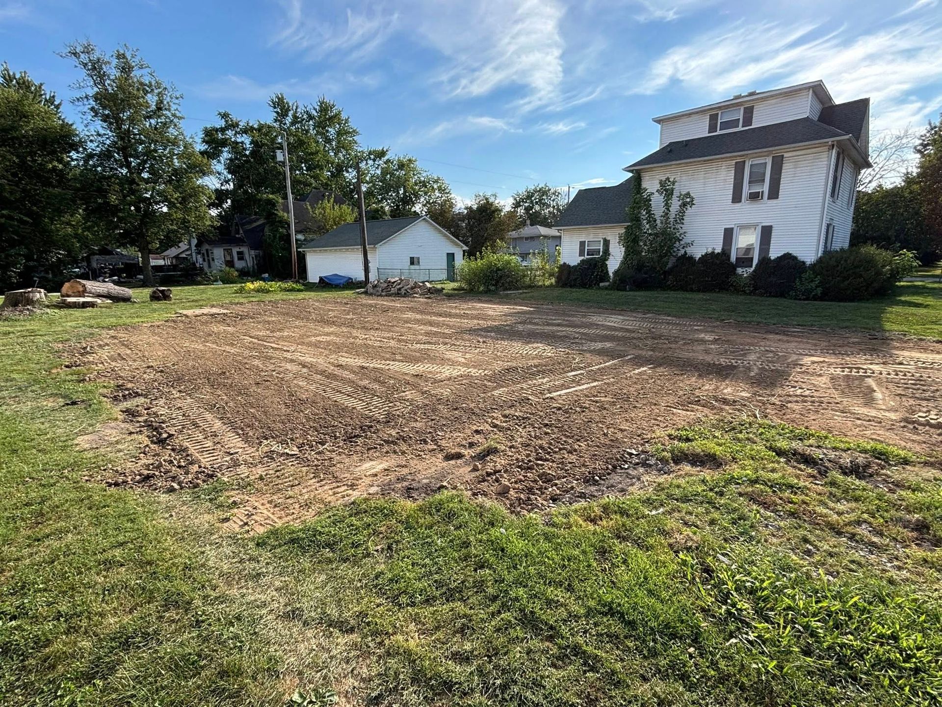 Cleared lot with brown soil, surrounded by green grass, houses in the background. Blue sky.