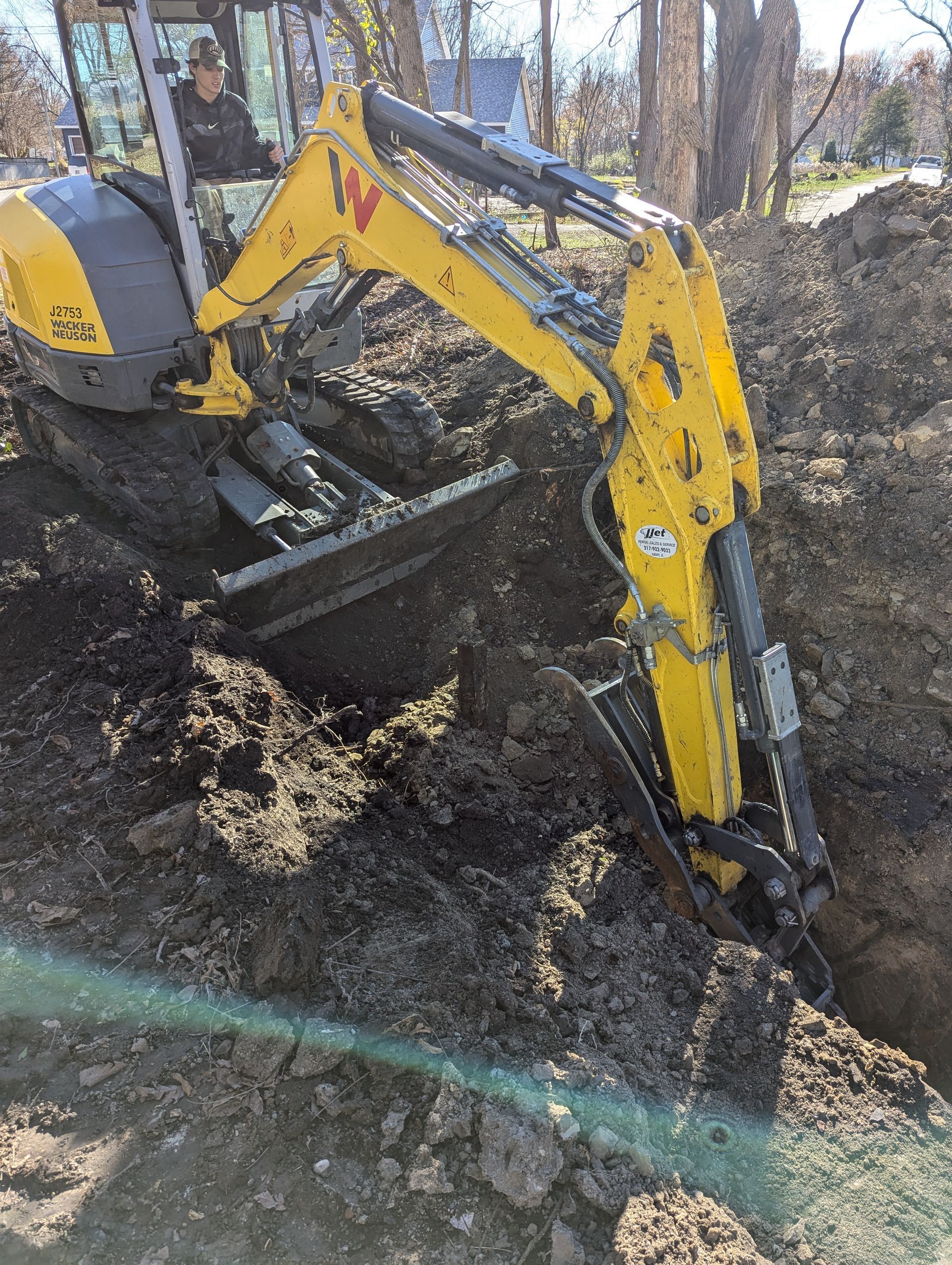 Yellow excavator digging a trench in brown soil outdoors. A person operates the machinery.