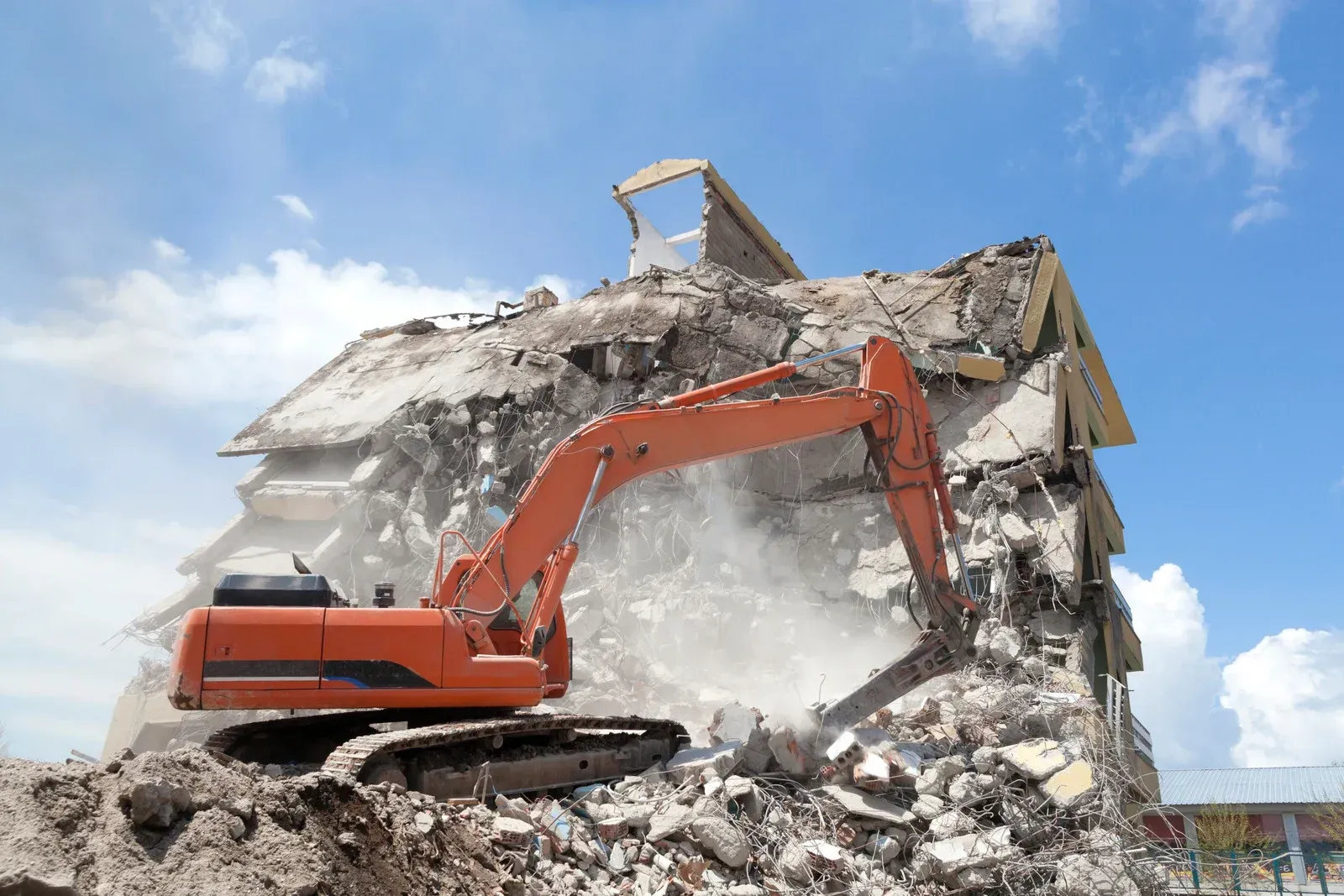 An orange excavator demolishes a building against a blue sky with clouds.