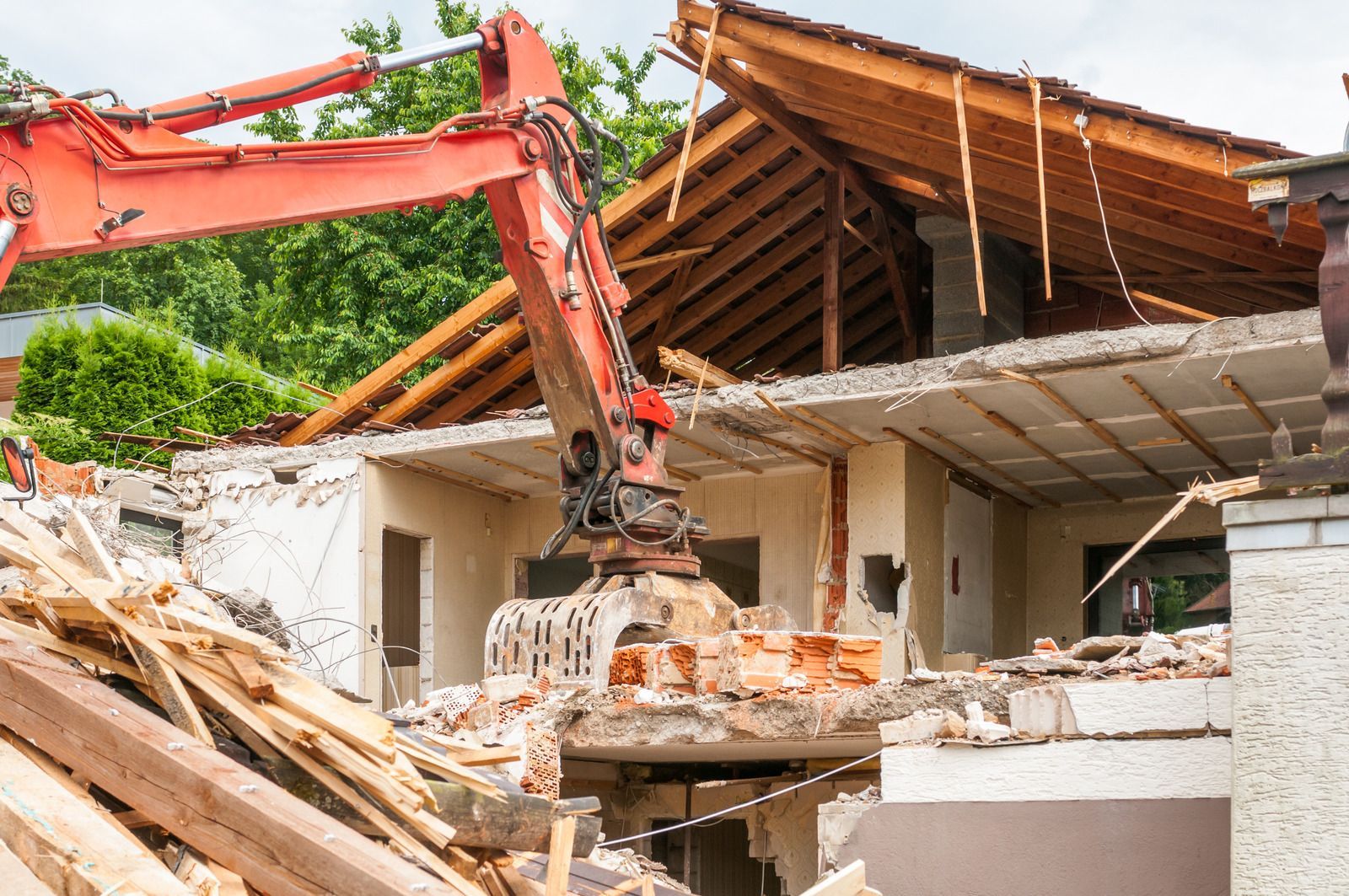 Red excavator demolishing a house; roof and walls being torn down in daytime.