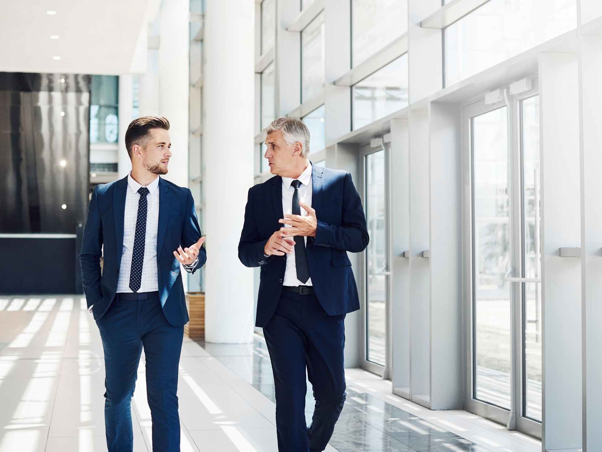 Two men in suits and ties are walking down a hallway and talking.
