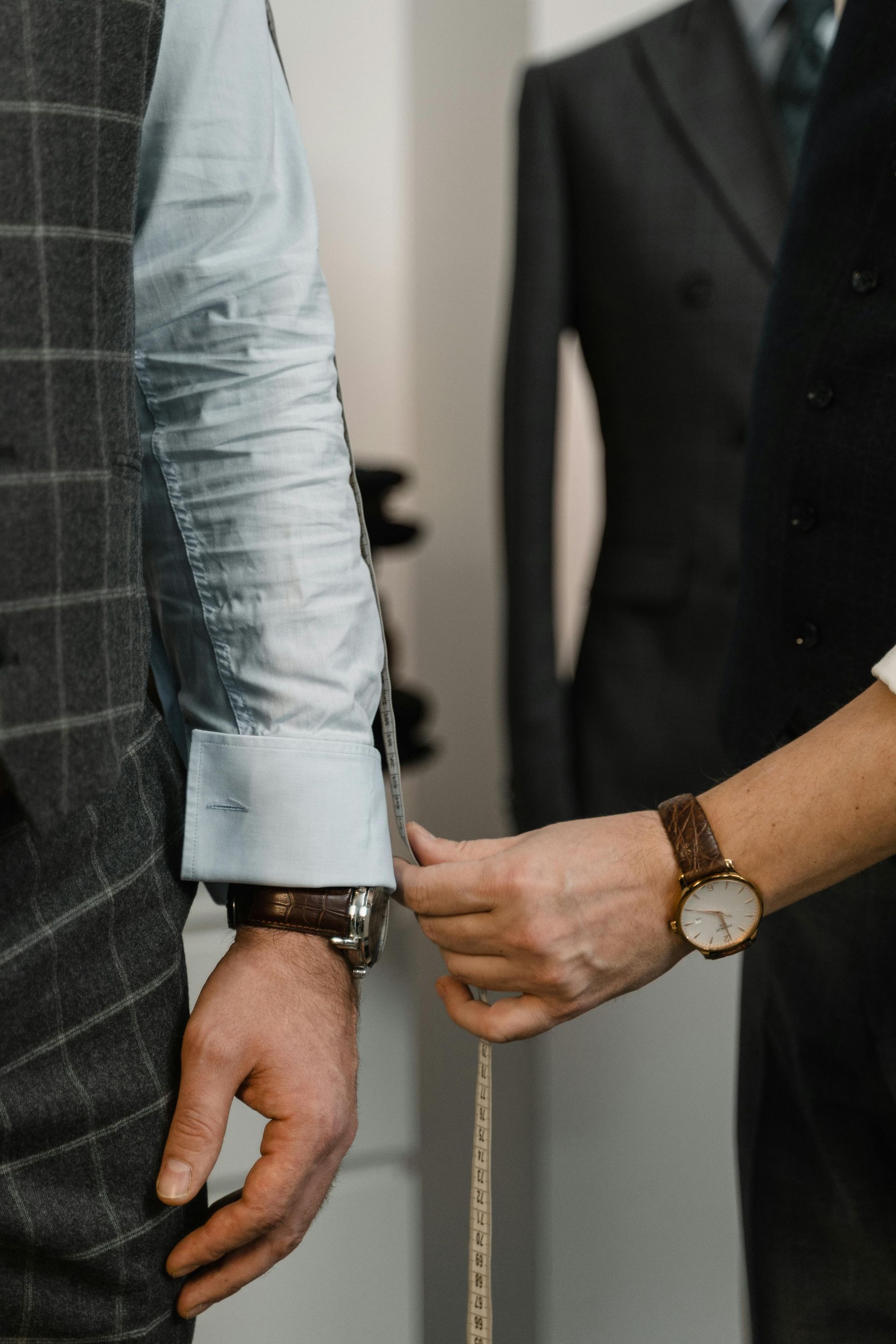 Tailor measuring a client's wrist for a suit fitting, with a suit jacket on a mannequin in the background.
