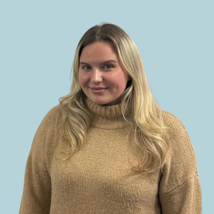Blonde woman with long hair in a tan sweater smiles at the camera against a light blue background.